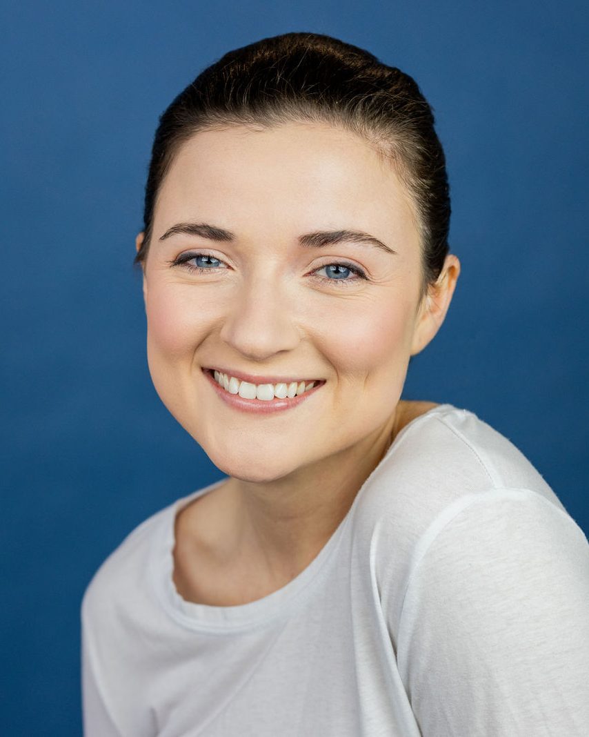 Actor headshot of a smiling woman in a white top against a deep blue backdrop, photographed by TM Headshots