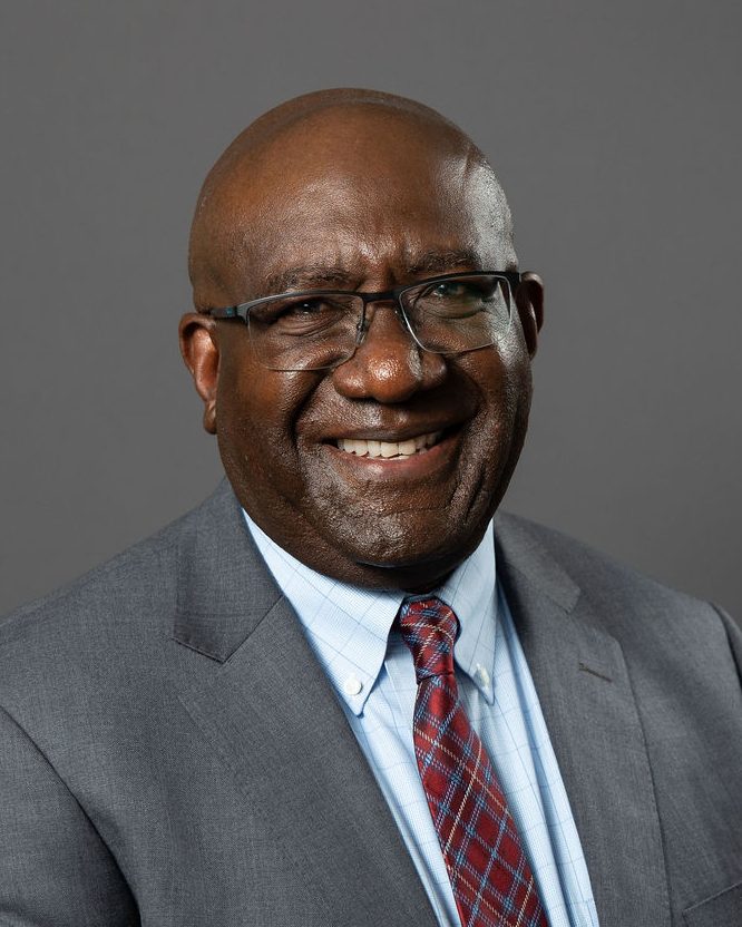 Corporate headshot of a smiling man in glasses and a gray suit against a dark gray backdrop, photographed by TM Headshots