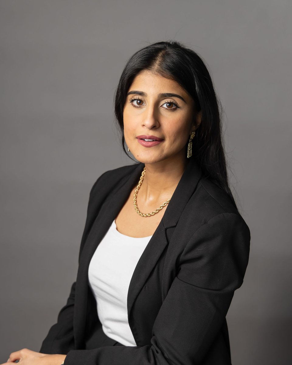 Editorial portrait of a woman in a black blazer and gold necklace seated against a gray backdrop, photographed by TM Headshots