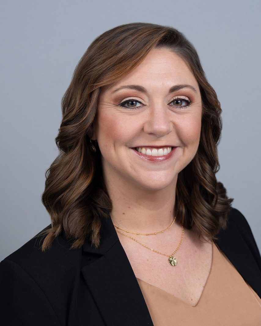 Professional headshot of a smiling woman with wavy brown hair in a black blazer against a blue-gray backdrop, photographed by TM Headshots
